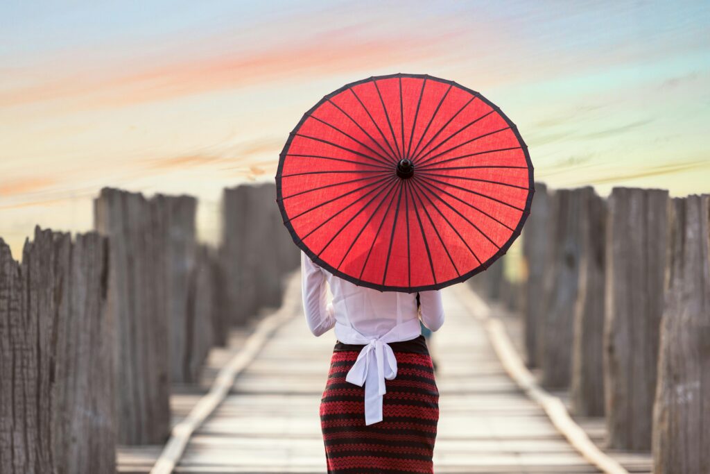 Woman with red umbrella walking on a wooden bridge at sunset.