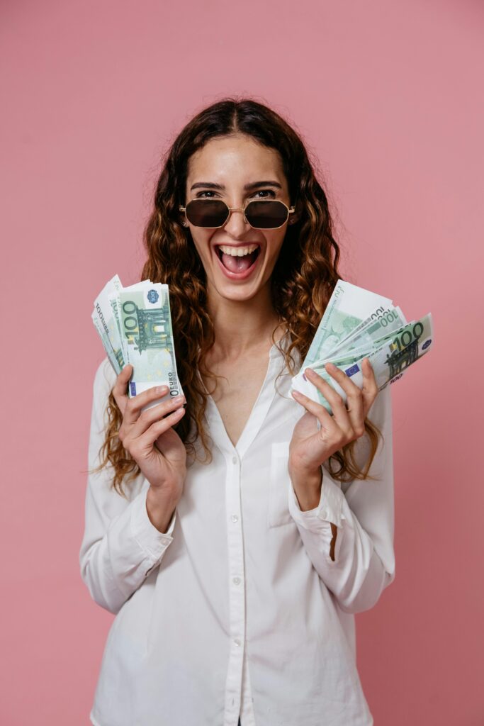 Smiling woman wearing sunglasses holding euro bills on a pink background.