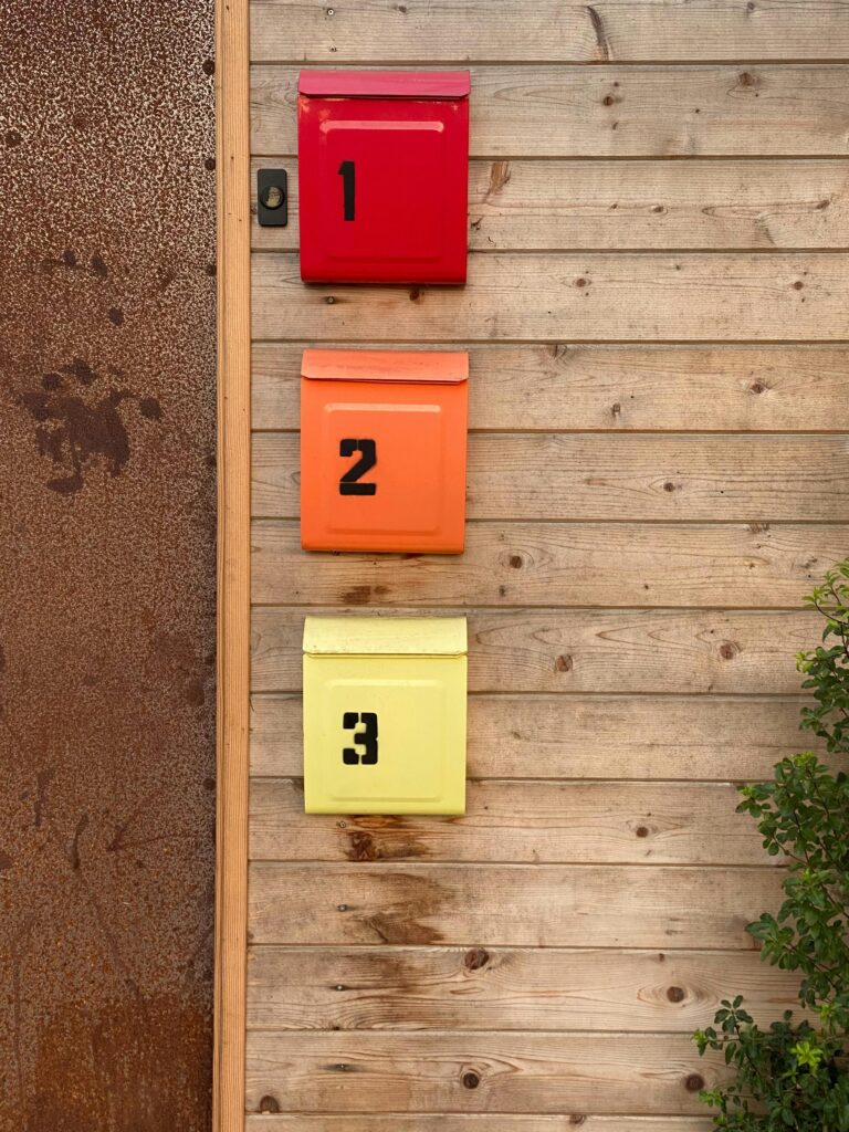 Colorful mailboxes with black numbers hanging on wooden wall near green plant and door in daylight